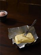 A pat of butter partially wrapped in foil with a butter knife in the middle sits on a dark wooden table with a small bowl of pink salt in the background.